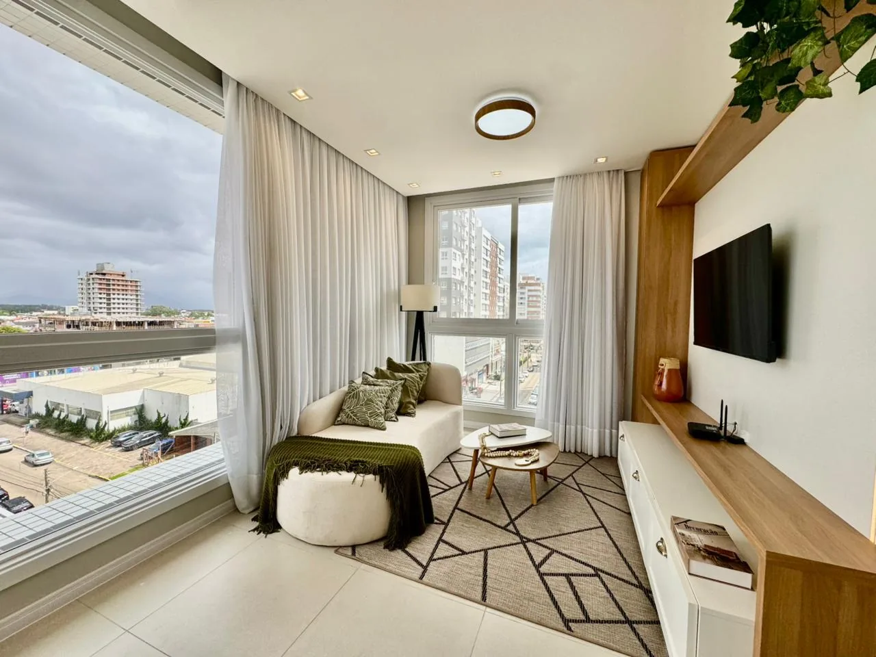Sala de estar com sofá claro, puff, tapete geométrico, TV em parede com moldura de madeira e janelas amplas com vista da cidade