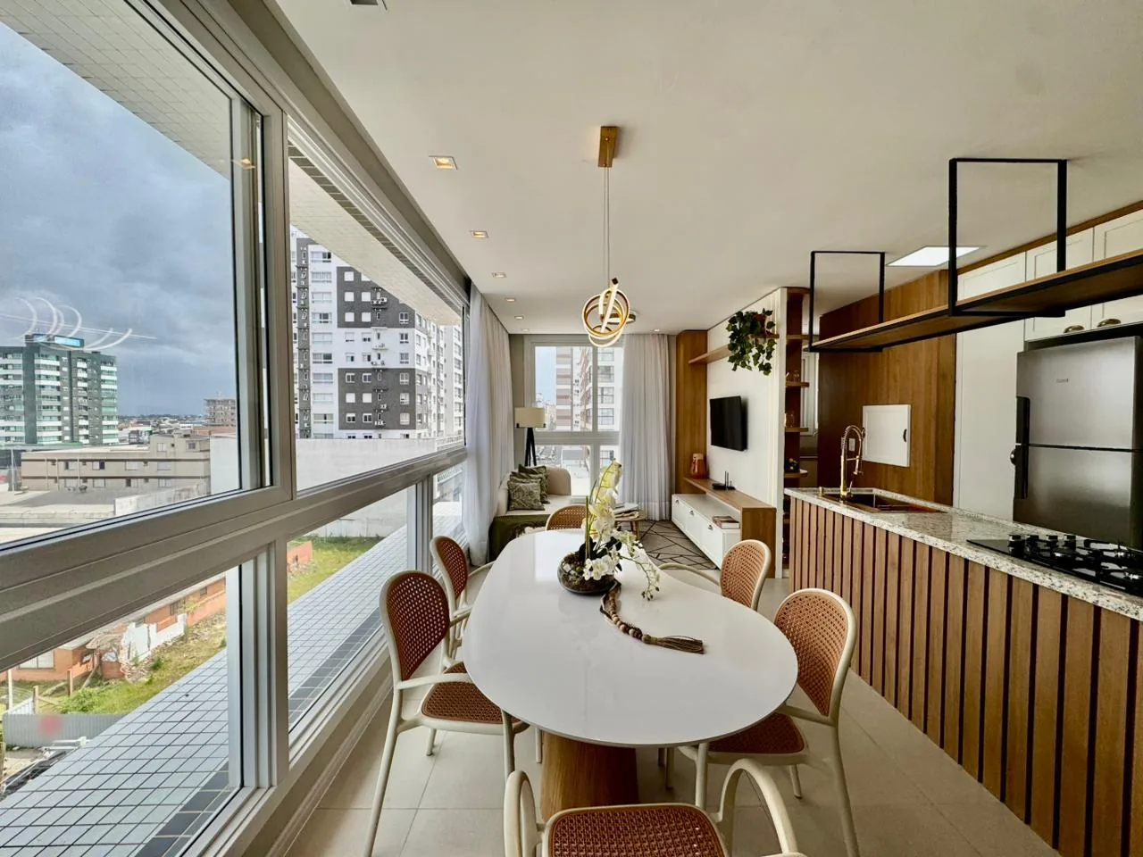 Mesa de jantar oval junto às janelas, ao lado da ilha da cozinha com cooktop, e sala de estar ao fundo