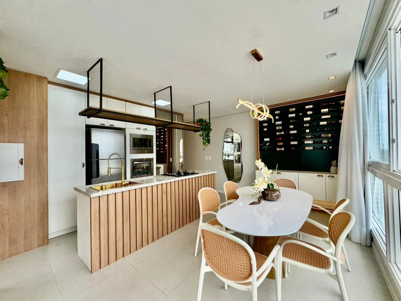 Cozinha com ilha ripada, torneira gourmet dourada, torre com micro-ondas e forno e mesa de jantar em frente à adega