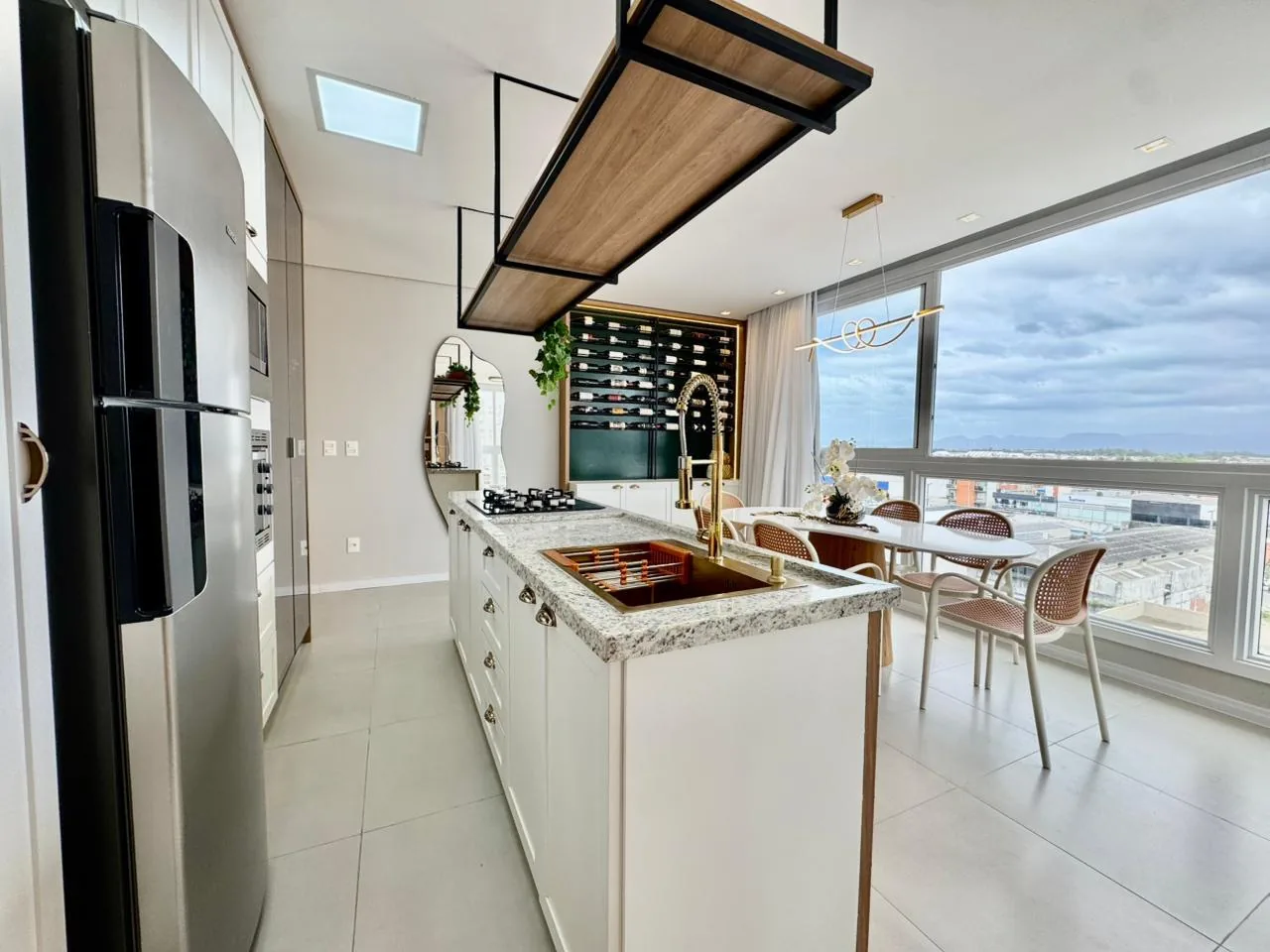 Ilha da cozinha com bancada de granito, cooktop e cuba cor cobre, geladeira inox e mesa de jantar junto às janelas
