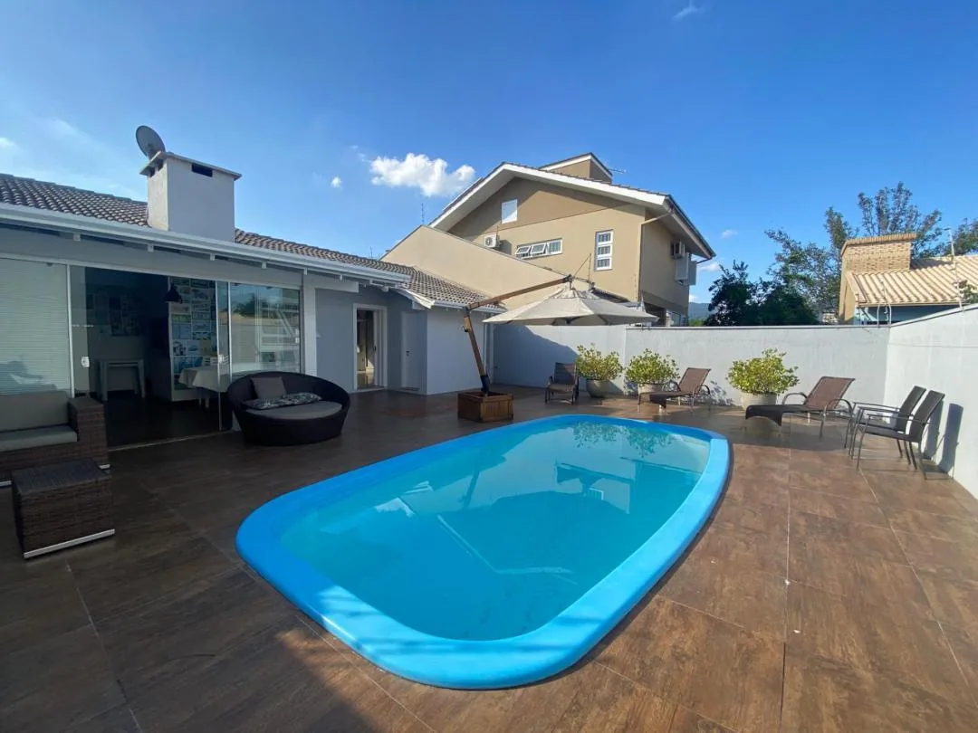 Piscina azul em piso amadeirado, com espreguiçadeiras, ombrelone e área gourmet envidraçada ao fundo