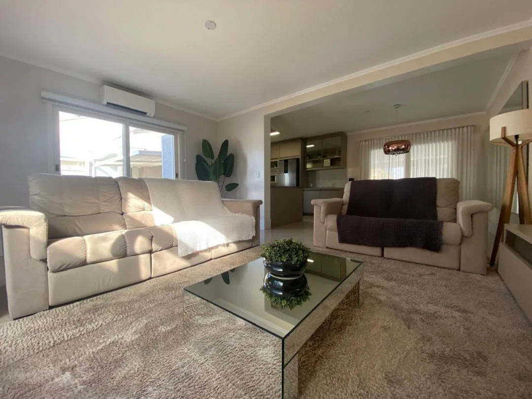 Sala de estar com dois sofás claros, tapete felpudo, mesa de centro espelhada e cozinha integrada ao fundo