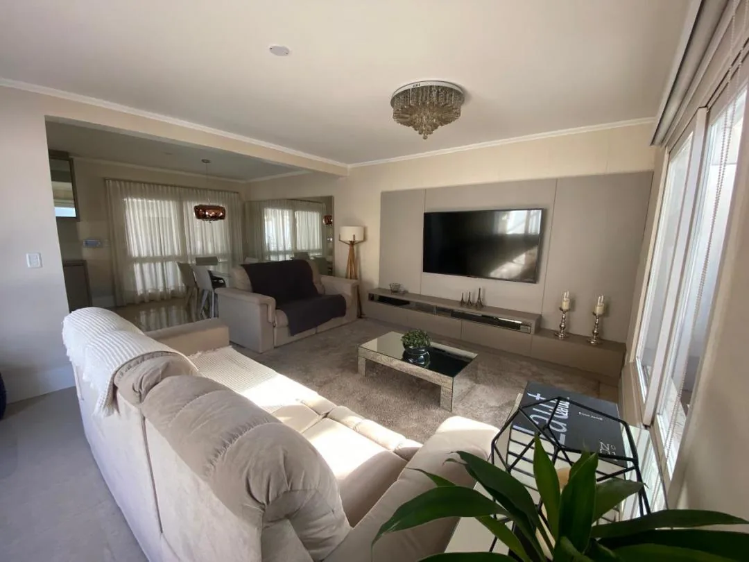 Sala com sofá bege, painel de TV com rack, lustre de cristal e jantar integrado ao fundo