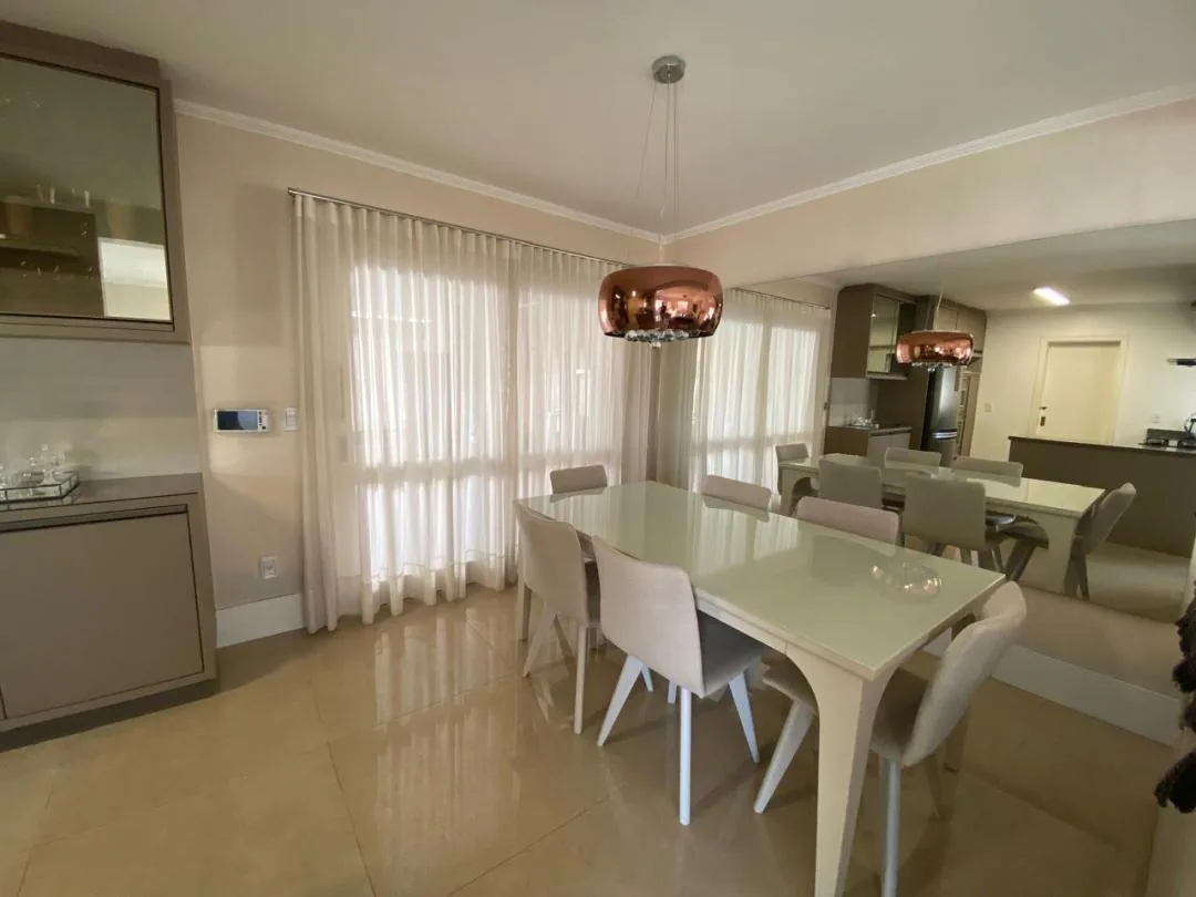 Sala de jantar com mesa de tampo de vidro, cadeiras estofadas claras, pendente acobreado e cortinas de voil