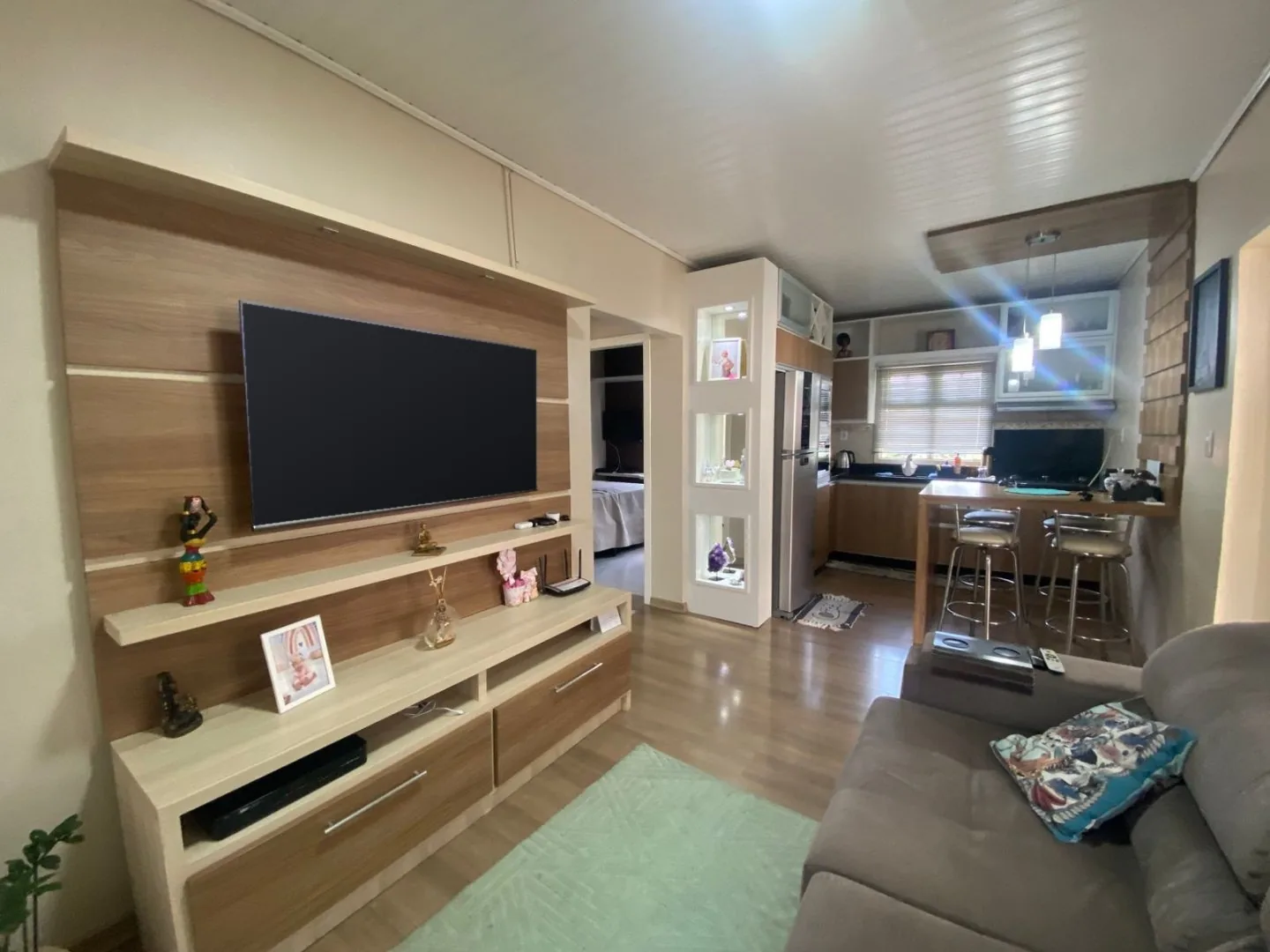 Sala de estar com painel de TV em madeira, sofá cinza e passagem aberta para a cozinha com balcão e banquetas