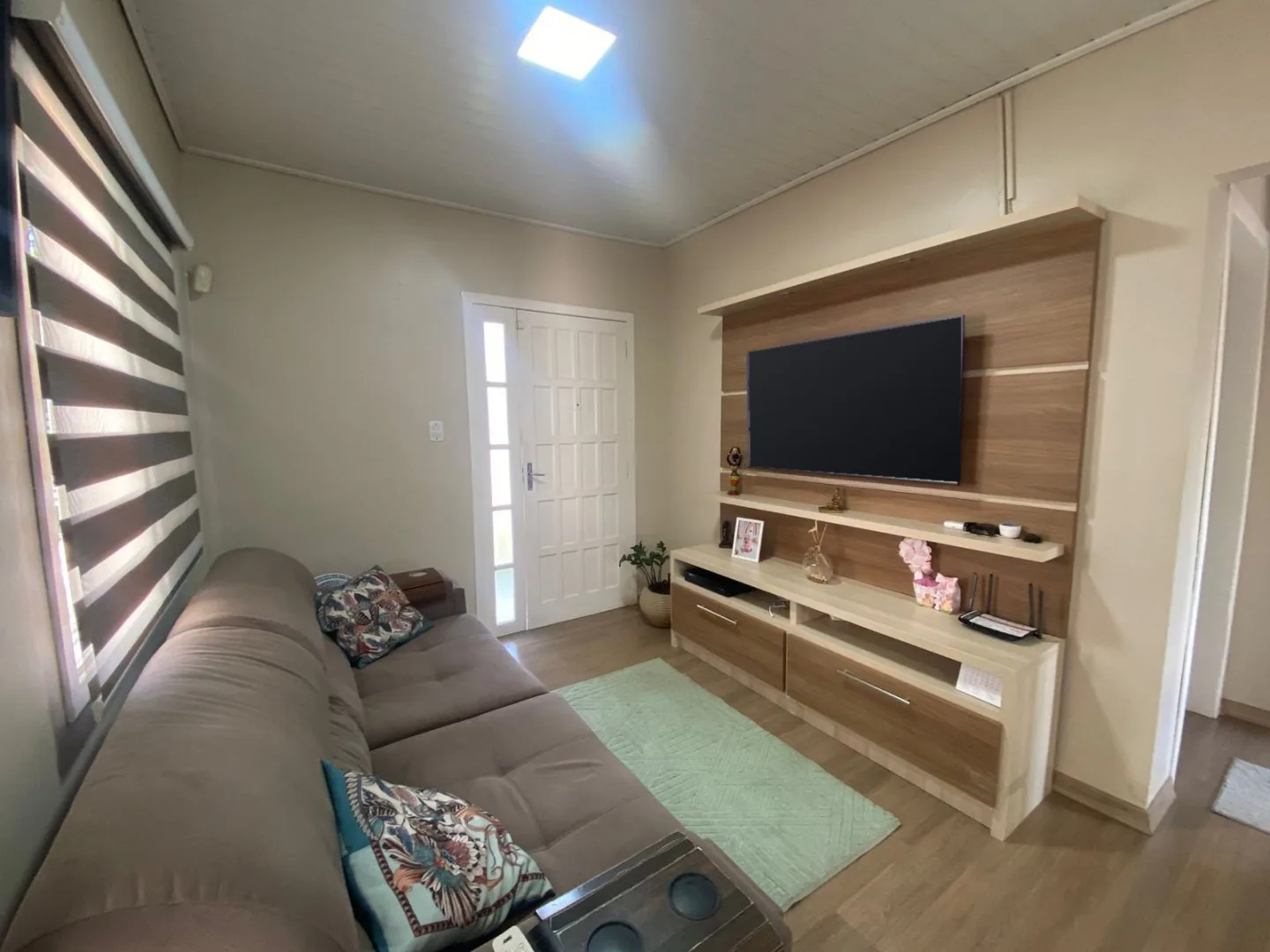 Sala com sofá cinza, cortina listrada, tapete verde-claro e painel de TV em madeira ao lado da porta de entrada branca