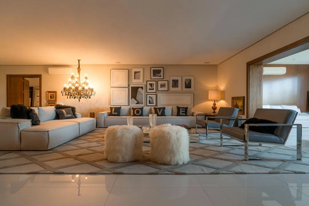 Sala de estar com sofá em L, dois pufes de pelo branco, poltronas de couro cinza e lustre de cristal aceso