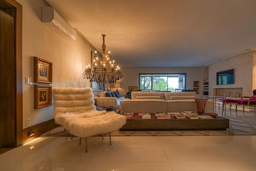Sala com poltrona de pelo branco, lustre de cristal, mesa de centro com livros e parede de pedra com TV ao fundo