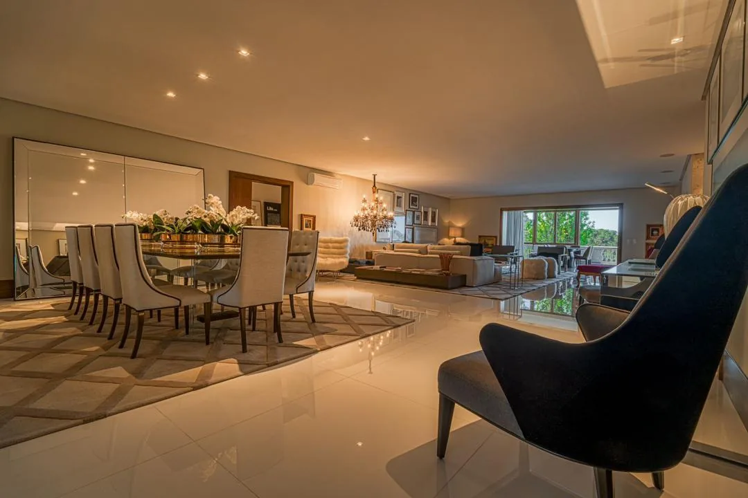 Sala de jantar com mesa de cadeiras estofadas claras, arranjo de orquídeas e grande espelho, integrada ao living com janela ao fundo
