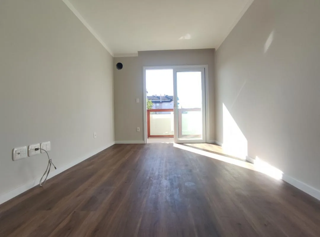 Sala vazia com piso vinílico amadeirado, paredes cinza-claro, tomadas na parede lateral e porta de vidro de correr para a sacada