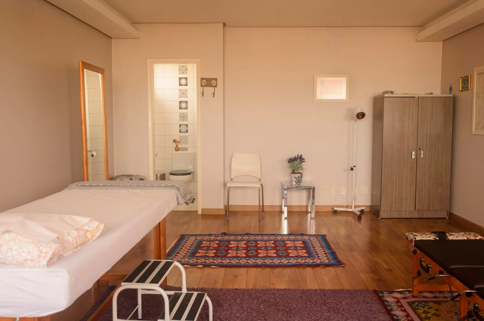 Sala com piso amadeirado, armário de duas portas, cadeira branca e banheiro com azulejos decorados ao fundo