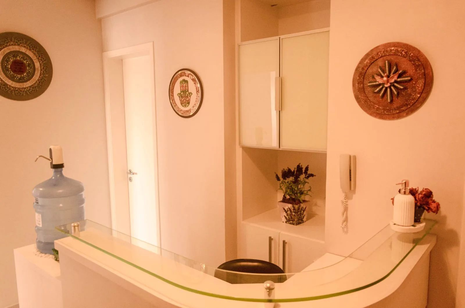 Balcão da recepção com tampo de vidro, armário branco embutido, interfone e porta de acesso