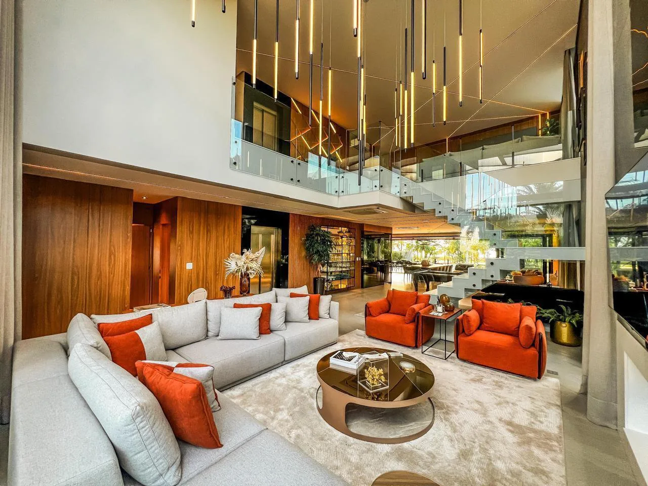 Sala de estar com pé-direito duplo, sofá cinza em L, poltronas laranja, pendentes lineares dourados e mezanino com guarda-corpo de vidro