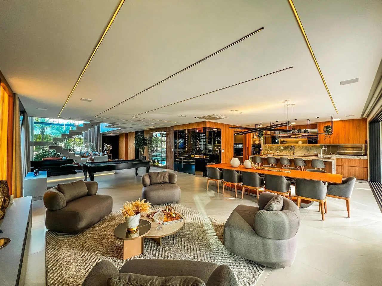 Living térreo integrado com poltronas arredondadas cinza, mesa de sinuca, mesa de jantar longa e cozinha com ilha ao fundo