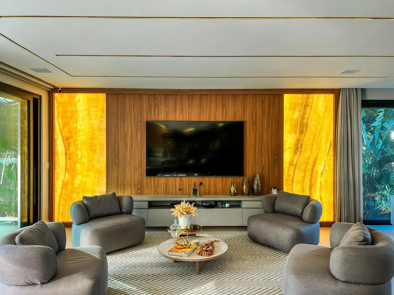 Sala de TV com painel de madeira entre dois painéis de ônix iluminados e quatro poltronas cinza