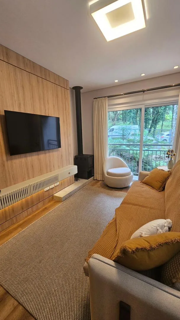 Sala com painel de madeira, TV na parede, lareira a lenha preta, poltrona bege e porta de vidro com árvores do lado de fora
