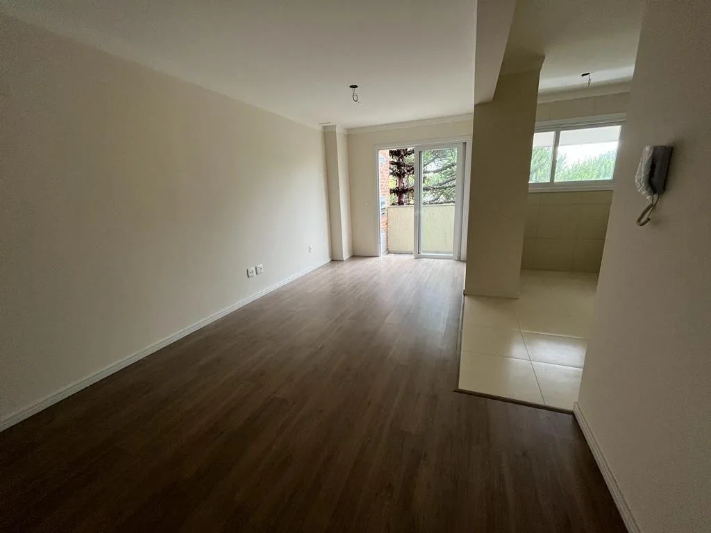 Sala ampla e vazia com piso laminado escuro, cozinha integrada à direita e porta de vidro para a sacada ao fundo