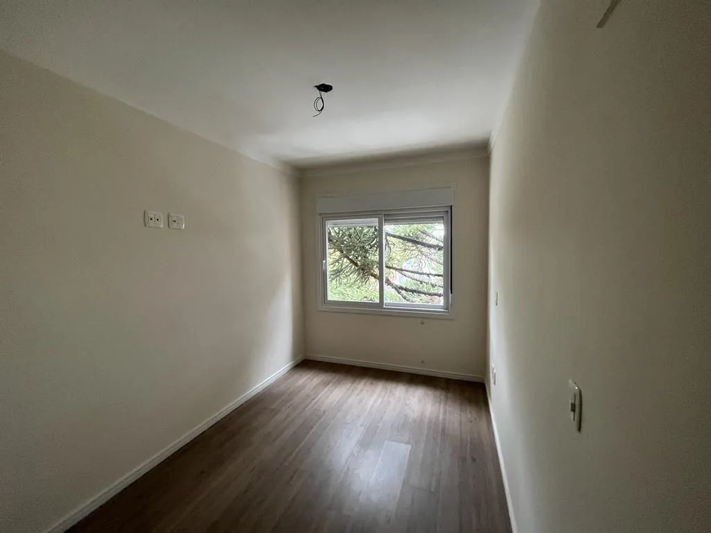 Quarto com piso laminado, paredes claras e janela de correr com persiana voltada para uma araucária