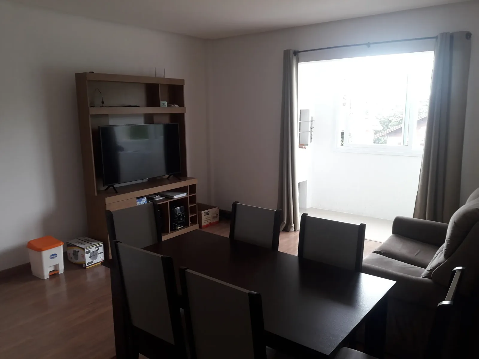 Sala com estante e TV, mesa de jantar escura e janela ampla com cortinas