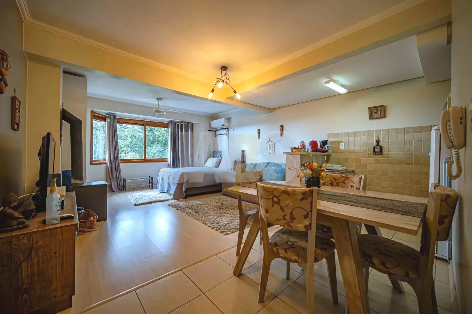 Sala com mesa de jantar e cama ao fundo, janelas para o verde